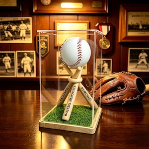 Op de afbeelding: Een witte honkbal met rode stiksels rust op een standaard van drie houten miniatuur honkbalknuppels in een doorzichtige acryl vitrinekast. De knuppels zijn gegraveerd met de tekst BASEBALL MEMORIES op een kunstgras ondergrond.