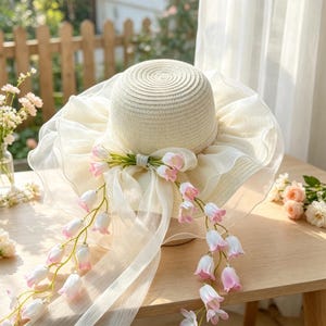 May include: A cream-colored straw hat featuring a wide, ruffled sheer organza brim. The hat is adorned with a central cream ribbon bow and two cascading floral vines with small, bell-shaped pink and white flowers hanging down the front.