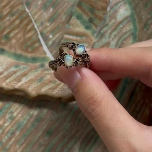 May include: Two stacked rings featuring oval-shaped iridescent opals set in dark, oxidized metal bands. The metal bands display intricate, raised floral or leaf-like engravings that wrap around the circumference of each ring.
