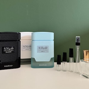 Op de afbeelding: Drie rechthoekige parfumflesjes met getextureerde bovenkanten in zwart, wit en lichtblauw, gelabeld Shiyaaka Shadow, Snow en Sky van Khadlaj. Daarnaast staan vier kleinere doorzichtige glazen proefflesjes en één klein doorzichtig flesje.