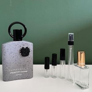 Op de afbeelding: Een grote grijze fles met zwarte spikkels, een zwarte ringdop en een zwart embleem, gelabeld Supremacy Collector's Edition Afnan. Daarnaast staan vier kleinere doorzichtige glazen verstuivers met zwarte doppen en een glazen fles met gouden dop.
