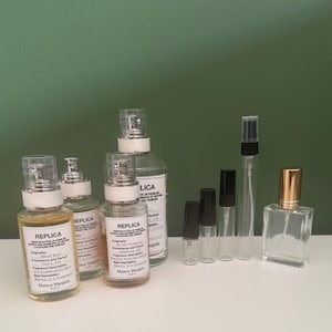 Op de afbeelding: Een verzameling parfumflesjes en verstuivers van helder glas. Drie grotere flessen hebben witte etiketten met zwarte tekst met de tekst REPLICA en Maison Margiela. Vier kleine slanke flesjes hebben zwarte doppen en een vierkante fles.