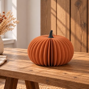 May include: A decorative pumpkin made of folded orange paper with a solid black stem. The pumpkin features a ribbed, accordion-style design that creates a rounded, symmetrical shape. It is a standalone object with a matte finish and clean lines.