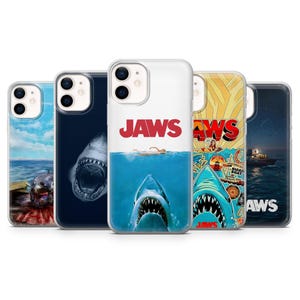 Op de afbeelding: Vijf doorzichtige smartphonehoesjes met diverse Jaws-filmgraphics. De ontwerpen tonen een haai die uit het water komt, een haaienbek op een donkere achtergrond, de iconische filmposter, een kleurrijke collage en een boot op de oceaan bij nacht.