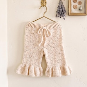 Puede incluir: Un pantalón de ganchillo en color beige claro con cintura acanalada, cordón ajustable y bajos con volantes acampanados. El pantalón cuelga de una percha de metal dorado, junto a un manojo de lavanda seca y un pequeño expositor enmarcado.