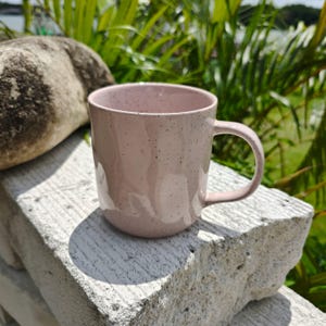 May include: A light pink ceramic mug with a speckled finish and a smooth, rounded body. The mug features a single curved handle on the right side and a wide, open top rim. The surface has a glossy texture with small dark flecks distributed throughout.