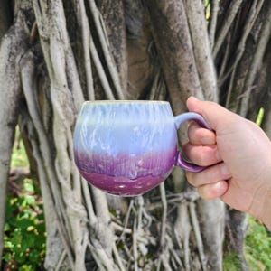 May include: A round ceramic mug featuring a smooth, glossy glaze that transitions from a pale, icy blue at the top to a deep, vibrant magenta at the base. The mug has a curved handle on the right side and a rounded, bulbous body shape.