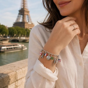 May include: A silver charm bracelet featuring various travel-themed pendants, including miniature flags of Canada, France, Brazil, and the United States, a detailed Eiffel Tower, a globe, a Statue of Liberty, and a small aeroplane charm.