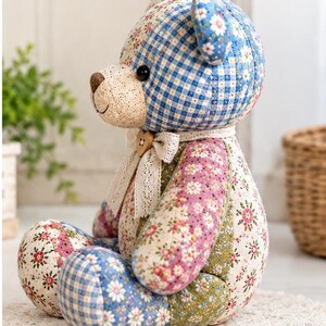 Pode incluir: Um urso de peluche feito de v&aacute;rios tecidos de patchwork com xadrez azul, padr&otilde;es florais em rosa, verde e branco, e um focinho cor de creme. O urso usa uma fita de renda &agrave; volta do pesco&ccedil;o com um pequeno bot&atilde;o de madeira em forma de cora&ccedil;&atilde;o.