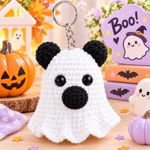 May include: A white crocheted ghost keychain featuring two round black ears, a round black nose, and two large black plastic safety eyes. The ghost is attached to a silver-toned metal split ring by a short chain, creating a small hanging accessory.