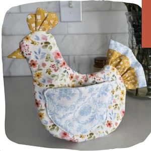 May include: A fabric chicken-shaped kitchen accessory featuring a white base with a multi-colored floral print in shades of pink, yellow, and green. It has a solid mustard yellow comb and beak, and a light blue patterned fabric wing pocket.