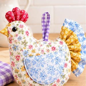 Könnte beinhalten: Ein ausgestopftes Stoffhuhn-Ornament mit einem cremefarbenen Körper mit buntem Blumenmuster, einem roten Blumenkamm, einem gelben Schnabel, einem schwarzen Auge, einem blauen Blumenflügel-Aufnäher, einer violetten Vichy-Karo-Schlaufe und einem gelb-blauen Rüschenschwanz.