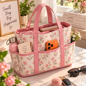 Peut inclure: Un sac fourre-tout rectangulaire en toile avec un fond crème, un motif floral rose et des bordures roses unies. Le sac possède deux anses, plusieurs poches extérieures et une ouverture supérieure. Il contient une gourde rose, un carnet et un téléphone.