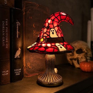 May include: A stained glass table lamp shaped like a witch hat with a red and black mosaic pattern featuring white ghost figures. The lamp has a dark, textured, fluted base and emits a warm glow from within the hat structure.