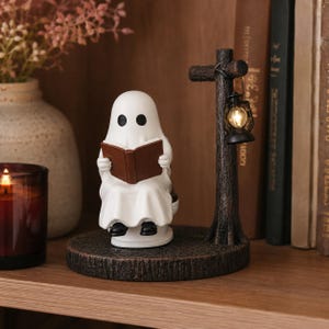 May include: A white ghost figurine sitting on a circular base, reading a brown book. Next to the ghost is a dark brown textured post featuring a small hanging lantern that emits a warm light. The entire structure is crafted with a detailed finish.