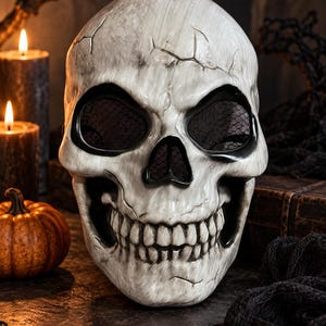 Peut inclure: Un masque de crâne blanc avec une texture vieillie et craquelée, présentant des détails noirs autour des orbites et de la cavité nasale. Les ouvertures des yeux sont recouvertes d'un filet noir fin et la mâchoire affiche un large sourire.