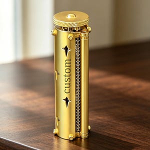 May include: A cylindrical gold-coloured metal rolling machine featuring a textured top knob, a vertical row of star-shaped cutouts, and the word custom engraved in black lettering on the side, with a visible internal metal mesh rolling mechanism.