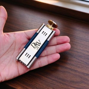 May include: A rectangular silver-toned metal lighter featuring a blue ribbed grip, a brass-colored ignition wheel, and a black engraved design with the letters D and W above the text Taylor and Jacob, accented by three small horizontal vents.
