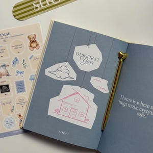 Könnte beinhalten: Ein offenes Erinnerungsbuch mit blauen Seiten, dem Text Our First Year und einer rosa Strichzeichnung eines Hauses. Ein goldener Stift mit herzförmigem Aufsatz liegt auf der Seite. Ein Bogen mit Aufklebern und ein gestreiftes Lesezeichen liegen bei.