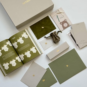 Op de afbeelding: Een babyherinneringsboekset in een grote beige doos, inclusief een groene fluwelen envelop, een klein zakje met trekkoord, een kaart met een berenillustratie met het label 1 Month, diverse beige en groene enveloppen en een boek met babykleding.