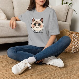 Può includere: Una t-shirt grigio melange a maniche corte con la grafica di un muso di gatto in stile cartoon con grandi occhi e la scritta "Peek-a-boo!" stampata in nero sotto. La maglietta è abbinata a jeans blu e scarpe da ginnastica bianche.