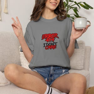 Może przedstawiać: Ciemnoszary t-shirt z krótkim rękawem z nadrukiem graficznym w kolorze czerwonym i czarnym z napisem ERROR 404 I DON'T CARE. Koszulka ma okrągły dekolt i luźny krój, prezentując wyrazistą czcionkę w stylu postarzanym, nadającą całości charakteru.