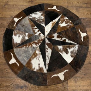 May include: A circular rug constructed from stitched cowhide patches in shades of brown, black, white, and gray. The pattern features a central star design with eight points, accented by four white cow skull silhouettes placed along the outer edge.