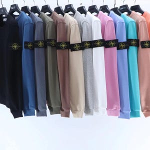 Può includere: Una collezione di felpe a maniche lunghe con girocollo appese a grucce nere, presentate in uno spettro di colori tra cui nero, blu, grigio, oliva, beige, bianco, rosa, viola, turchese e arancione, ciascuna con una toppa a bussola sulla manica.