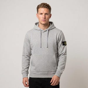 Puede incluir: Una sudadera con capucha y manga larga en color gris claro, con un bolsillo tipo canguro frontal, capucha con cordón ajustable y un parche circular negro y amarillo con un diseño de brújula en la manga izquierda. Tejido de punto liso.
