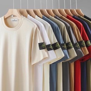 Peut inclure: Une collection de t-shirts à manches courtes et col rond suspendus sur des cintres en bois. Chaque t-shirt présente une couleur unie et un écusson rectangulaire noir sur la manche gauche avec un logo boussole jaune et vert. Les couleurs incluent crème, blanc, bleu, gris, beige, rouge, marine, olive et noir.