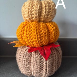 May include: A stacked crochet pumpkin decoration featuring three tiers in tan, orange, and beige yarn. The top pumpkin is topped with a small brown twig stem. A single red maple leaf and a yellow maple leaf are tucked between the bottom two tiers.