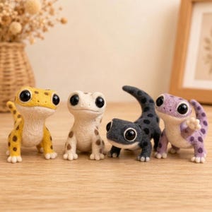 May include: Four needle-felted lizard figurines with large black eyes. The first is yellow with brown spots, the second is cream with brown spots, the third is dark grey with black spots, and the fourth is light purple with dark purple spots.