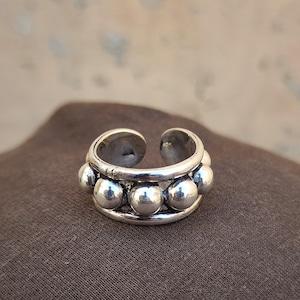 May include: A silver-toned open ring featuring a central band decorated with a series of smooth, raised spherical beads. The metal has a polished finish with darkened recessed areas between the spheres, creating a distinct contrast across the band.