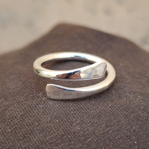 Op de afbeelding: Een gepolijste zilverkleurige bypass-ring met een glad, open ontwerp. De ring bestaat uit één doorlopend stuk metaal dat om de vinger wikkelt, met twee taps toelopende, platte uiteinden die elkaar licht overlappen voor een strakke uitstraling.