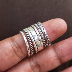 Puede incluir: Un conjunto de seis anillos de tono plateado con texturas y diseños variados, que incluyen una banda martillada, una banda de cuerda trenzada, una banda lisa pulida, una banda fina simple, una banda de cuentas y una banda con patrón floral.