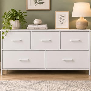 May include: A white dresser featuring five drawers with horizontal white handles. The top row consists of three smaller drawers, while the bottom row contains two larger drawers. The unit is supported by a simple frame with four legs.