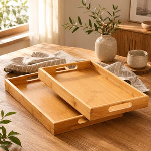 May include: Two rectangular bamboo serving trays with cut-out handles on the short sides. The trays feature a natural light wood grain finish and are stacked slightly offset, revealing the smooth interior surfaces and sturdy raised edges.