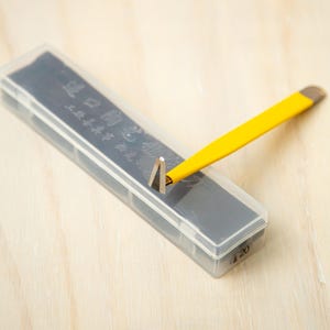 Op de afbeelding: Een precisiegereedschap met een gele handgreep en een metalen driehoekige punt, rustend op een rechthoekige opbergdoos van doorzichtig plastic. De doos bevat een donkergrijze inzet met tekst en het getal 20 in de rechterbenedenhoek.