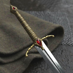 May include: A dagger with a metallic brass-toned hilt featuring a textured, rope-like grip and a pommel topped with a small red gemstone. The crossguard is adorned with a larger, marquise-cut red gemstone at its center, leading to a steel blade.