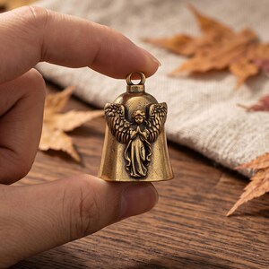 May include: A small, antique-style brass bell featuring a raised relief design of an angel with outstretched wings and hands pressed together in prayer. The bell has a smooth, conical body and a small circular loop at the top for hanging.