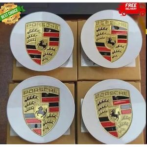 May include: A set of four circular white wheel center caps, each featuring a gold, red, and black Porsche crest emblem in the center. The crest displays the word PORSCHE at the top, a central horse, and horizontal red and black stripes.