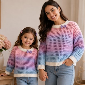 May include: Two matching long-sleeve sweaters featuring a waffle-knit texture with a gradient color pattern transitioning from pink to purple to blue. Each sweater has a white ribbed crew neckline, white cuffs, and a small purple bow detail.