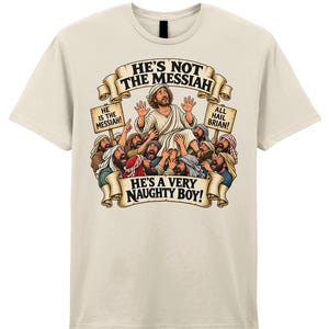 May include: A cream-coloured short-sleeve t-shirt featuring a graphic illustration of a crowd lifting a man. The text reads HE'S NOT THE MESSIAH, HE IS THE MESSIAH!, ALL HAIL BRIAN!, and HE'S A VERY NAUGHTY BOY! on beige banners and signs.