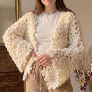 May include: A chunky, off-white, handmade crochet cardigan featuring a repeating floral motif throughout the knit. The garment has long, wide sleeves and an open front design. The text "riff.label" and "Handmade Chunky Crochet Cardigan | Pakistani Design" is visible.