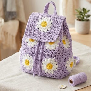 May include: A lavender crochet backpack featuring a pattern of white and yellow daisy motifs. The bag has a top handle, a flap closure, and a drawstring cord made of the same lavender yarn. The construction consists of joined square panels.