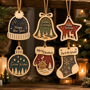 Op de afbeelding: Zes houten hangende ornamenten met touwtjes. De vormen omvatten een muts met "Happy New Year", een bel met "Season's Greetings", een ster met "Happy Holidays", een kerstbal met "Warm Wishes", een want met "Merry Christmas" en een kous met "Joy to the World".
