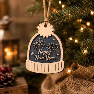 Op de afbeelding: Een houten ornament in de vorm van een wintermuts met een pompon, voorzien van een donkerblauw middenpaneel met gegraveerd vuurwerk, sterren en de tekst Happy New Year in wit schrift, opgehangen aan een bruin touwtje.