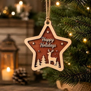 Op de afbeelding: Een stervormig houten ornament met een donkerbruin middenstuk waarin een hert en dennenbomen zijn uitgesneden. De tekst Happy Holidays is in het midden gegraveerd. Het ornament heeft een lichte houten rand en hangt aan een touwtje van jute.