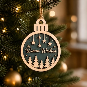 Op de afbeelding: Een ronde houten kerstornament met een donkergroene uitgesneden kern met de tekst Warm Wishes in een sierlijk lettertype. Het ontwerp bevat gelaagde houten dennenbomen aan de onderkant en hangende sterren, opgehangen aan een touwtje.