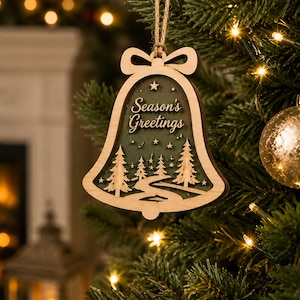 Op de afbeelding: Een klokvormige houten kerstornament met een gelaagd ontwerp en een donkergroene achtergrond. De uitsparing toont een kronkelend pad door een bos van dennenbomen onder een sterrenhemel, met de tekst Season's Greetings in een sierlijk lettertype.